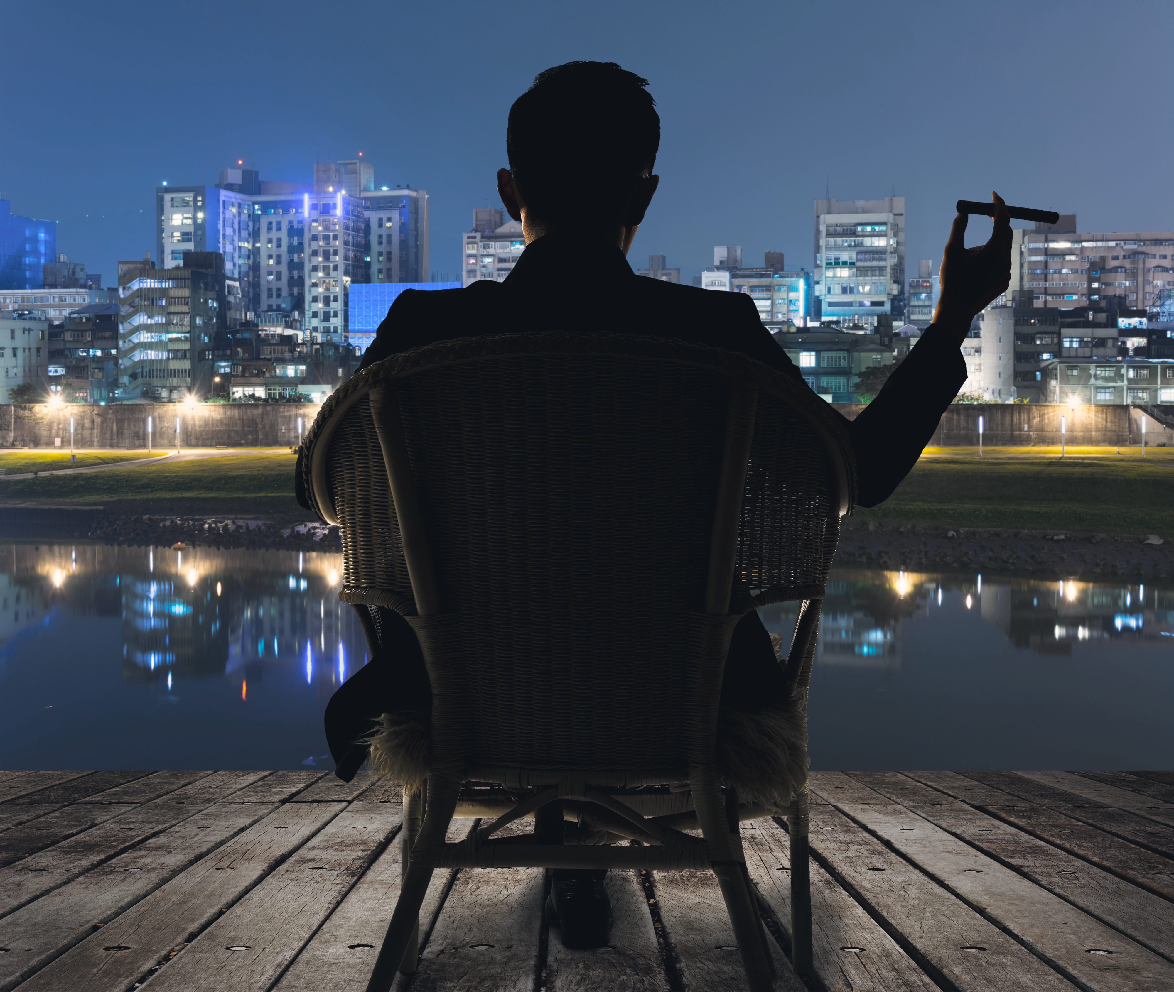 Silhouette of businessman sit on chair and hold a cigar and looking at the city in night.